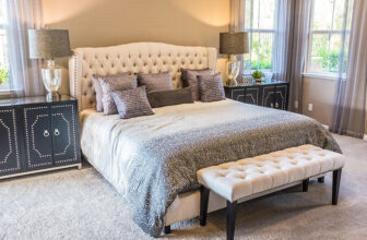 Przytulna sypialnia z pikowanym łóżkiem, neutralną pościelą i dekoracyjnymi poduszkami. Dwie czarne szafki nocne z lampami stoją po bokach łóżka, a pikowana ława stoi u stóp. Naturalne światło wpada przez okna z cienkimi firankami.