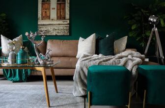 Stylowy salon z jasnobrązową sofą, zielonymi ścianami i turkusowymi pufami. Szklany stolik kawowy ma elementy dekoracyjne, a w pobliżu stoi lampa na trójnogu. Przytulny szary pled jest rozłożony na sofie. Nad sofą wisi oprawione zdjęcie drzwi.