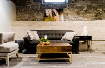 Przytulny salon z ciemnoszarą sofą ozdobioną poduszkami, drewnianym stolikiem kawowym z małą rośliną i świecą oraz krzesłem bocznym. Ściana ma odsłonięty kamień, a nad sofą zamontowano zdjęcie zieleni.
