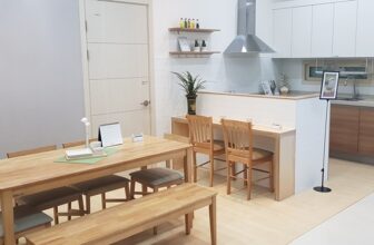 Nowoczesna kuchnia i jadalnia z meblami z jasnego drewna. Stół jadalny ma ławkę i krzesła, a kuchnia ma białe szafki, okap ze stali nierdzewnej i małe rośliny dekoracyjne. Na ścianie wisi oprawiony obraz.