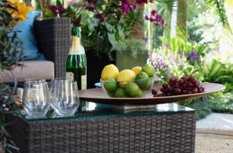 Wiklinowy stolik kawowy ze szklanym blatem eksponuje miskę cytryn i limonek, kiść winogron i butelkę szampana z kieliszkami. Tło przedstawia żywe fioletowe kwiaty i bujną zieloną roślinność na zewnątrz.